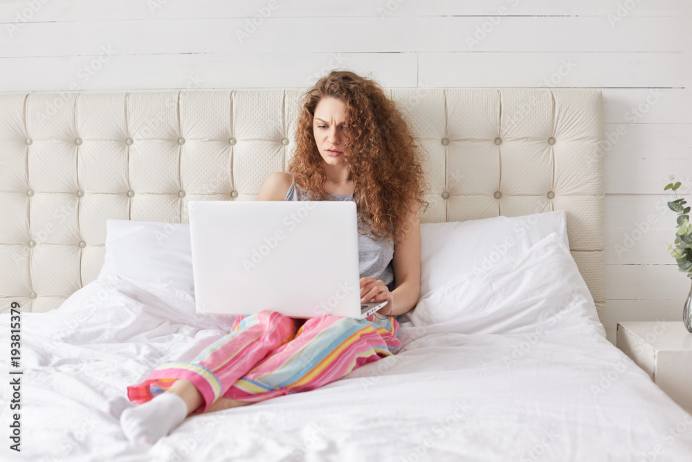 Serious young female freelancer uses laptop computer for remote job, relaxes in bed in home interior, dressed casually, searches information on web site, connected to wireless internet.