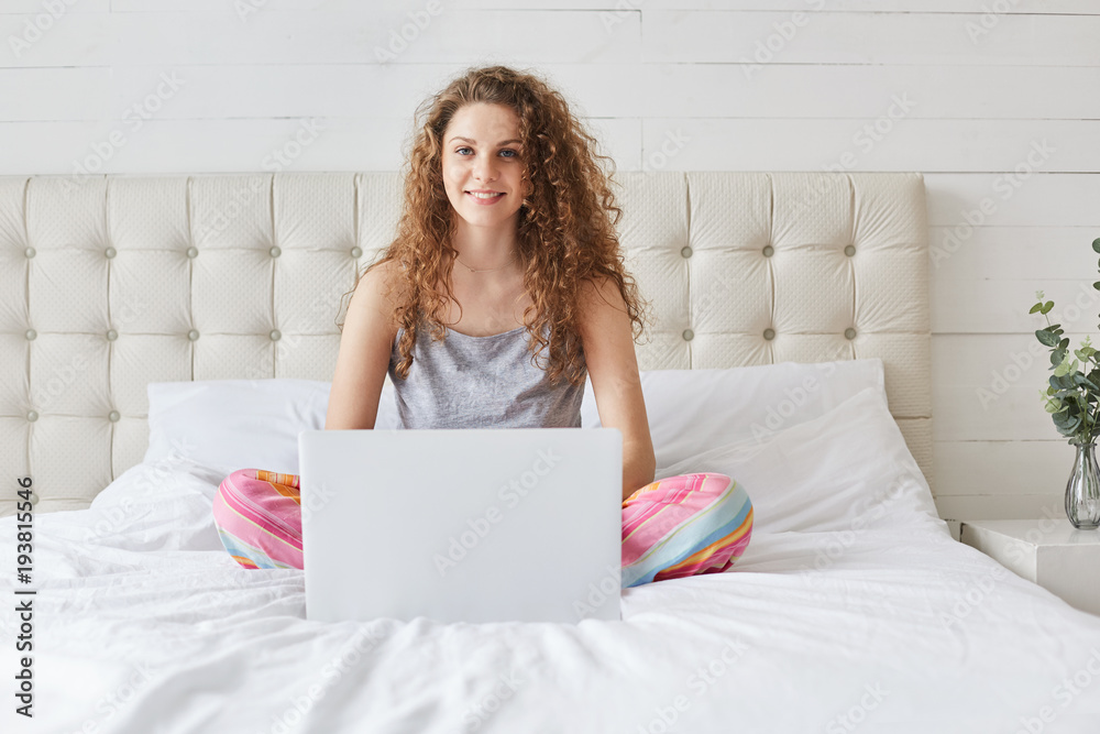 Positive beautiful female uses portable laptop computer for e learning, rests in domestic atmosphere at bedroom, dowloads files from networks or prepares for project. Woman in bedroom with device Positive beautiful female uses portable laptop computer for e learning, rests in domestic atmosphere at bedroom, dowloads files from networks or prepares for project. Woman in bedroom with device