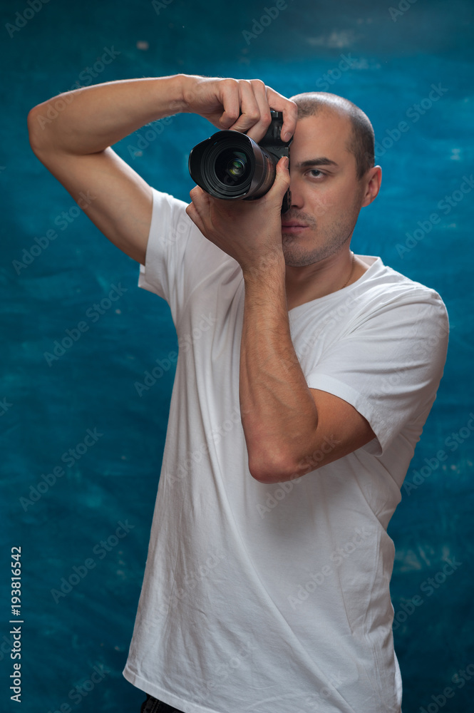 Obraz premium Smiling middle-aged man in white shirt with camera in his hands posing on blue background.