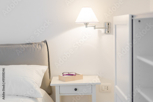 Detail of a bedroom. White interior. A lamp, a book and glasses on the nightstand. Relaxation concept