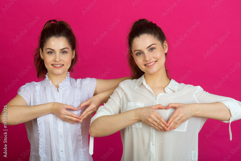 two beautiful young sisters twins in a good mood in white blouses on a pink background showing a hand gesture in the form of a heart two beautiful young sisters twins in a good mood in white blouses on a pink background showing a hand gesture in the form of a heart