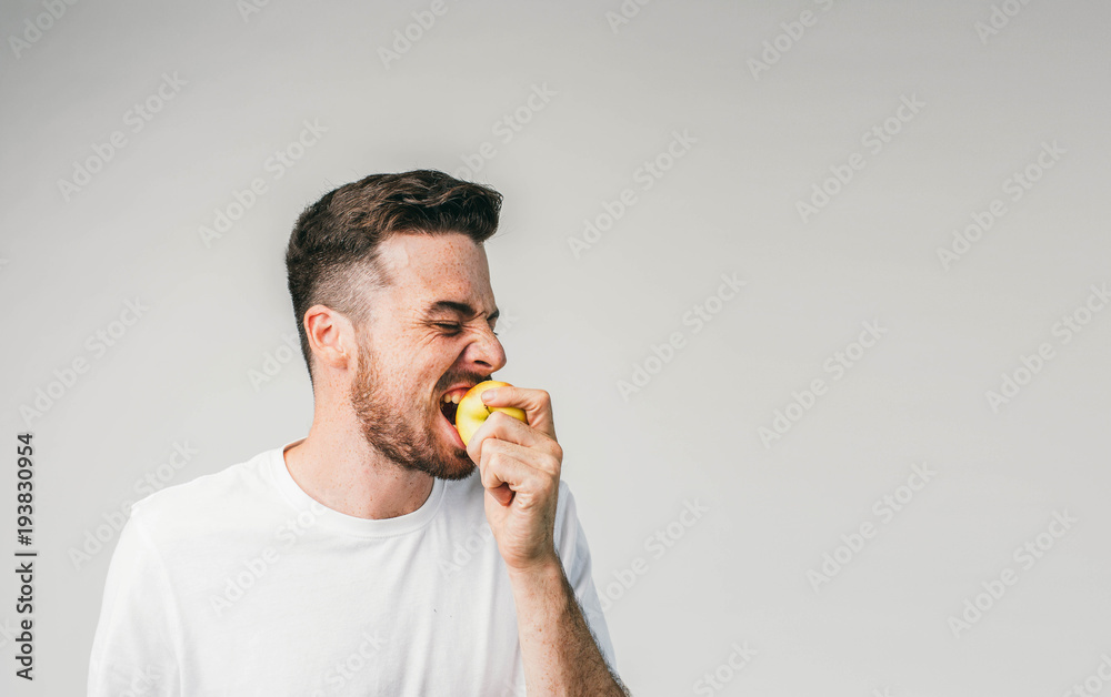 Guy is standing on the left side of the photo frame and biting an apple. This fruit is so sour that he cannot stop wiggling. Cut view.