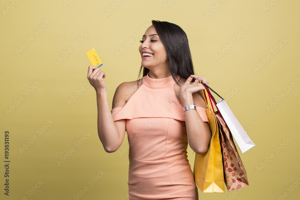 Portrait of a smiling pretty girl in dress with shopping bags holding credit card, isolated over yellow background