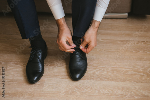 A man or businessman in a suit ties up shoelaces on black leather shoes on a parquet background