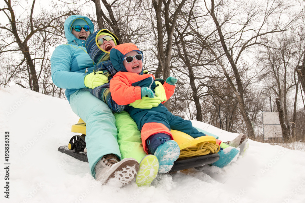 Happy mother with children in sunglasses and bright blue ski suit. Mother and boys are happy together. Kids ride a yellow sled in the forest with their mom. Guys are very happy to play outdoors
