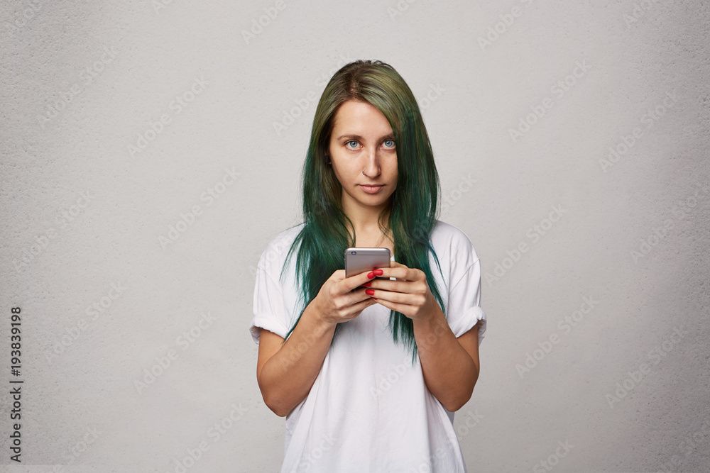 Young girl typing and looks directly into camera. Fashionable modern hairdo, dyed green hair. Isolated over white wall. Young girl typing and looks directly into camera. Fashionable modern hairdo, dyed green hair. Isolated over white wall.
