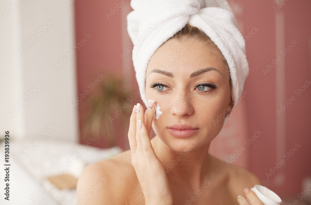 Young woman with flawless skin, applying moisturizing cream on her face. Photo of woman after bath in white bathrobe and towel. Skin care concept Young woman with flawless skin, applying moisturizing cream on her face. Photo of woman after bath in white bathrobe and towel. Skin care concept