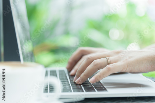 Asian woman hands and married ring has touching and typing on laptop computer with blurred coffee, computer and view outside window.