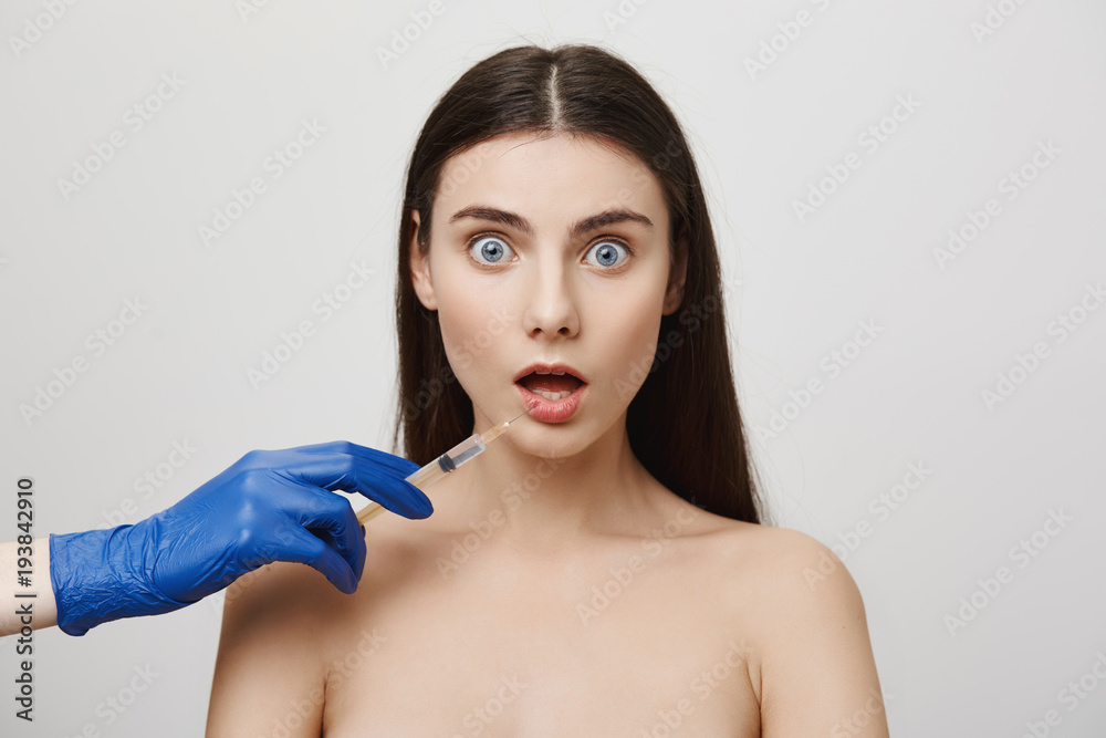 Obraz premium On her way to lip-augmentation. Portrait of nervous young woman receiving shot in lower lip from cosmetologist in medical glove, wanting to change her appearance and make face even more beautiful