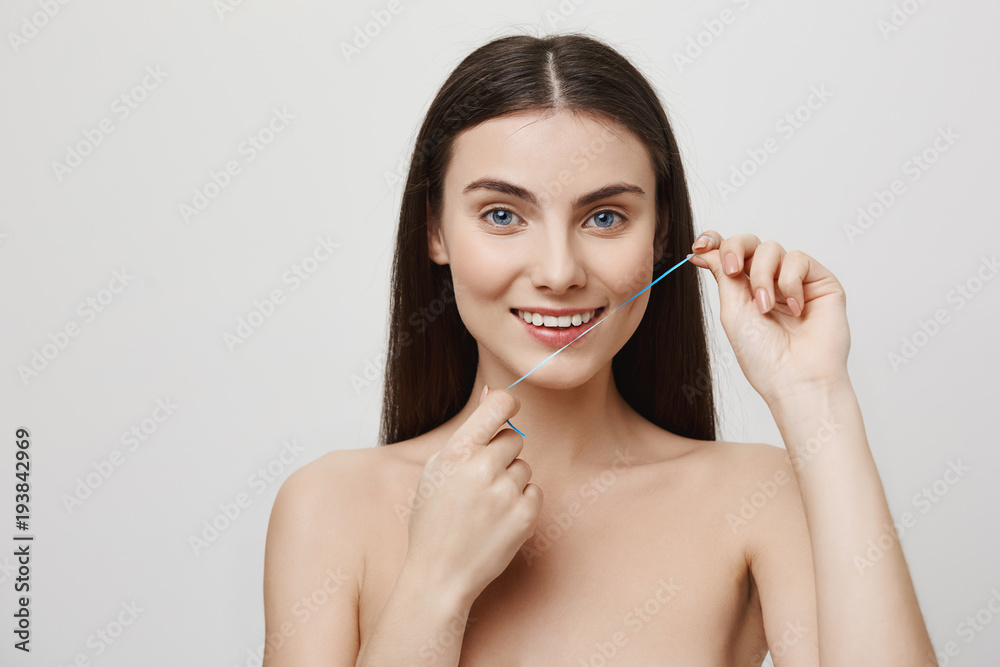 Taking care of perfect smile. Studio shot of beautiful woman getting treatment for teeth with dental floss, smiling while enjoying doing hygiene procedures. Girl makes sure she cleans everything