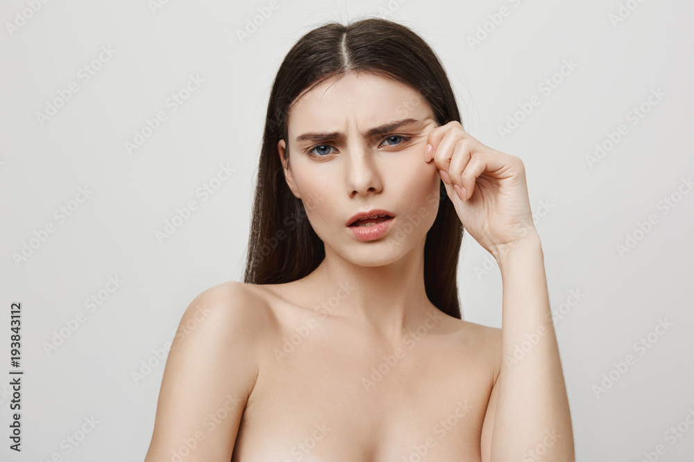 Wrinkles attacking her beautiful face. Studio shot of upset irritated young european woman, standing naked while pulling face near eye and showing aging results, being sad about it Wrinkles attacking her beautiful face. Studio shot of upset irritated young european woman, standing naked while pulling face near eye and showing aging results, being sad about it