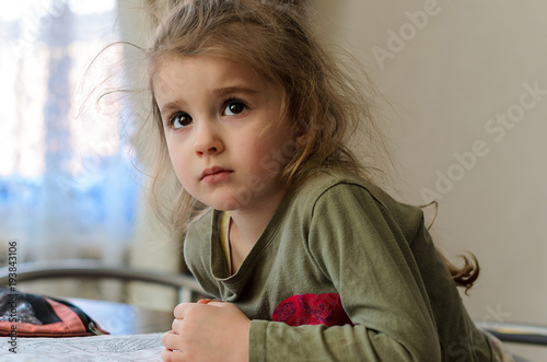 a little girl with expressive brown eyes sits at a wooden brown table and looks frightened up, next to her painting supplies