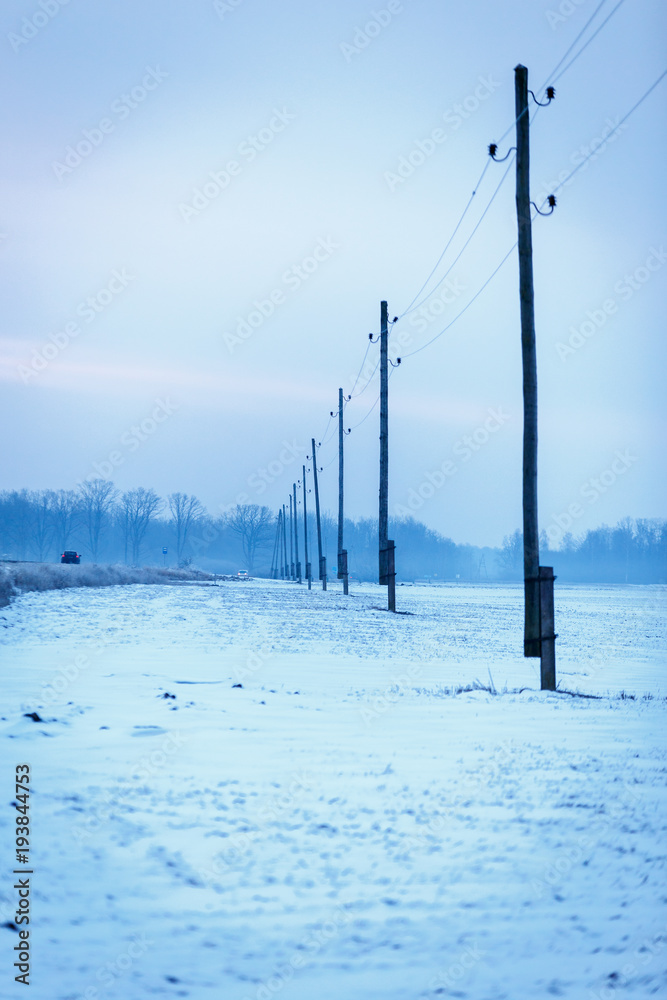 Early winter cold foggy morning - electricity poles row. Ground covers snow, along the way is a field and electricity poles row. visible in the distance trees, haze.