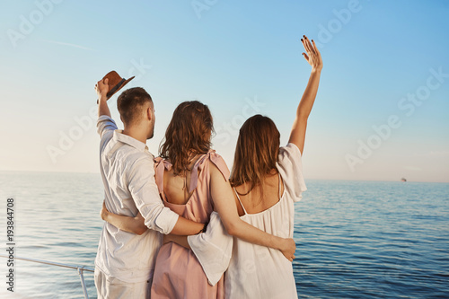 Back view of three best friends travelling by boat hugging and waving while looking at sea. People who are on luxury vacation say hi to ship crue that passes by yacht.