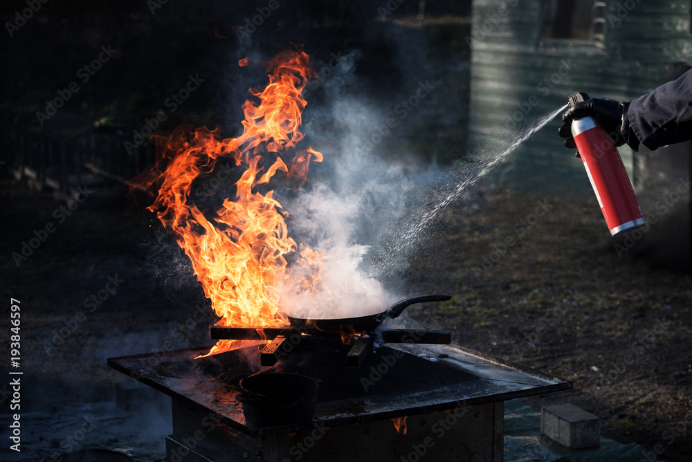 Obraz premium Man extinguishing the fire on an iron pan with foam from a spray can, demonstration during a fire department training, dark background with copy space