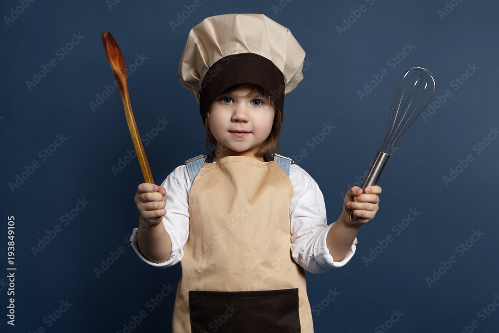 Naklejka premium Children, cooking, cuisine and food concept. Picture of funny little girl wearing chef uniform standing at blank wall, holding rolling pin in one hand and two wooden spoons in other, smiling at camera