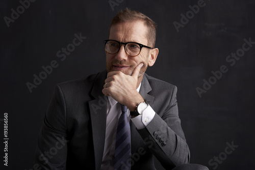 Thinking businessman portrait. Studio shot of wrinkled face business man wearing suit and looking thoughtful while sitting at dark background. Professional man wearing suit and tie. 