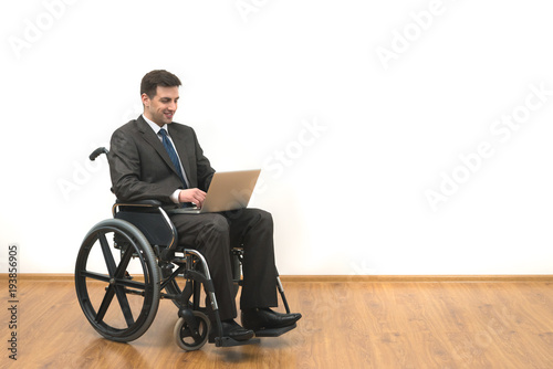 The smile man in a wheelchair sitting with a laptop on a white wall background