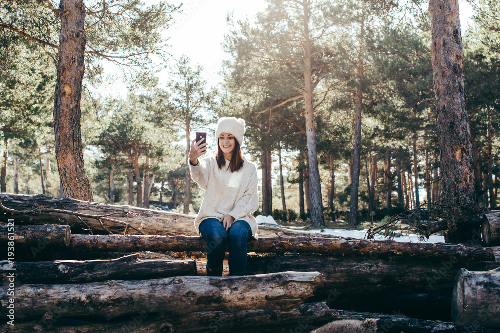 Fototapeta premium Young and pretty woman using her mobile phone outdoors, having fun taking herself pictures on a winter day with seated in a wooden trunk. Lifestyle.