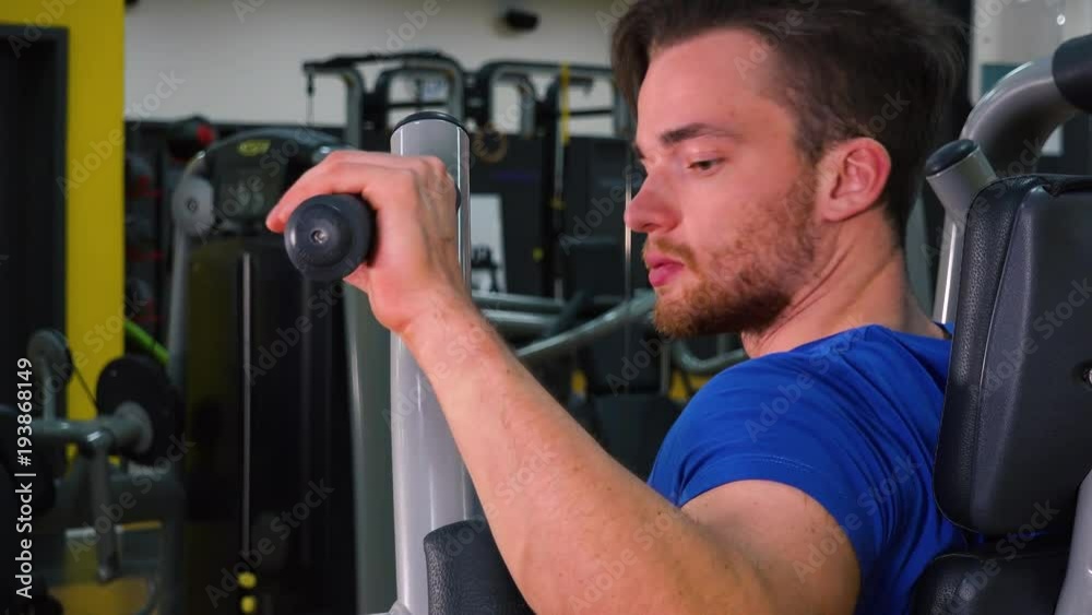 A young fit man walks to a machine in a gym and starts working out - closeup