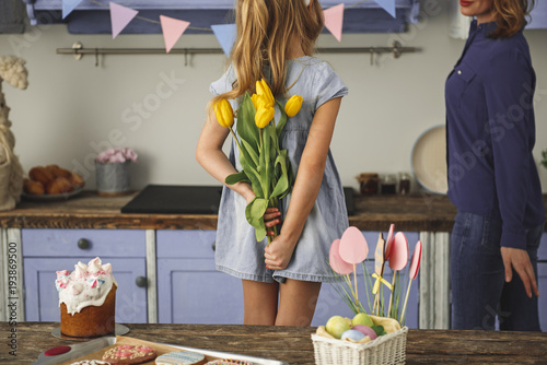 Young girl standing in kitchen and hiding bunch of yellow tulips behind her back while looking at her mother walking by. Copy space in left side