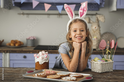 Portrait of beautiful young girl in country style kitchen sitting at table with easter cake, cookies and basket with colorful eggs inside. She is looking at camera with smile. Copy space in left side