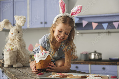 Cheerful little girl in cute bunny ears headband sitting on kitchen bar table with easter cake and looking at cookies lying in front of her. Rabbit toy on background