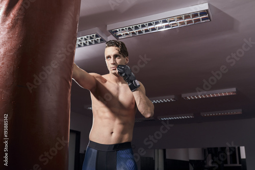 one boxer portrait, adult man, shirtless posing, puching boxing bag, indoors room. wearing boxing gloves, tights. photo