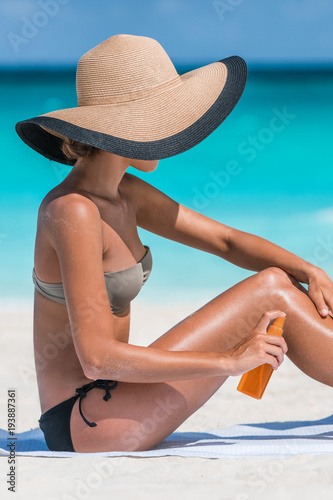 Sexy bikini woman putting suntan lotion oil on tanned skin. Girl tourist applying sunblock cream or mosquito repellent on body holding bottle. Model wearing sun hat.