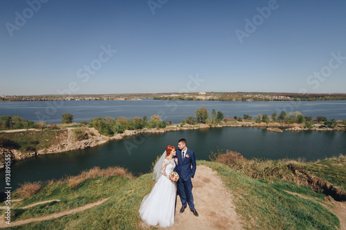 Beautiful young bride and groom stand on a cliff in the background of the river and gently embrace each other. The red-haired bride. Summer wedding.