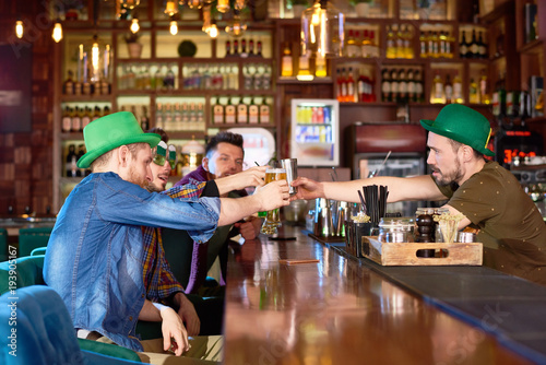 Group of friends clinking beer glasses together while gathered together in modern pub in order to celebrate St. Patricks Day, some of them wearing green bowler hats