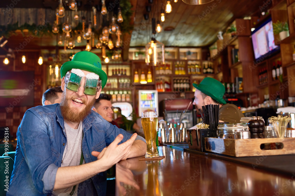 Portrait of joyful red-haired man wearing funny party glasses and green bowler hat looking at camera with toothy smile while sitting at bar counter, group of friends behind him