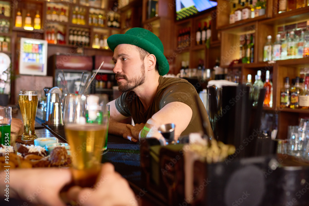 Profile view of handsome bearded barman wearing green bowler hat leaning on bar counter while entertaining visitors with small talk, portrait shot
