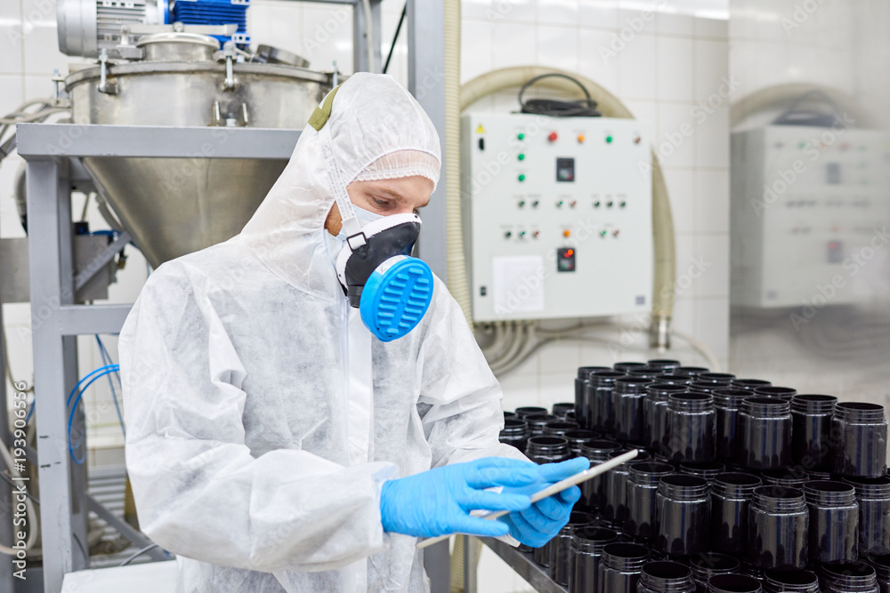 Confident young worker wearing coverall and respirator using digital tablet while taking inventory in warehouse of pharmaceutical factory Confident young worker wearing coverall and respirator using digital tablet while taking inventory in warehouse of pharmaceutical factory
