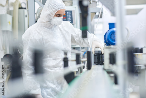 Young man wearing coverall and safety mask working on production line of modern pharmaceutical factory, portrait shot photo