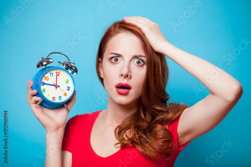 Young beautfiul redhead girl with alarm clock on blue background