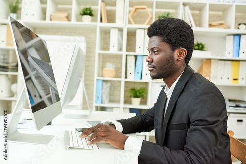 Profile view of confident African American manager sitting at desk and working on promising project with help of computer, interior of modern office on background