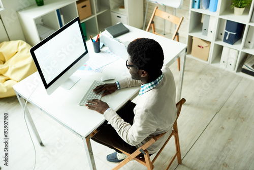 Obraz na plátně High angle view of hard-working African American entrepreneur sitting at office desk and preparing visual presentation for his business partners with help of modern computer
