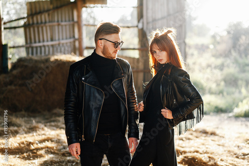 a bearded guy stands with his girl in black leather jackets in a barn