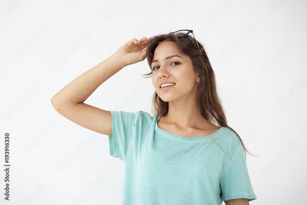 Optics, eyewear and style concept. Studio shot of charming smiling young mixed race woman with long brunette hair posing isolated wearing casual blue t-shirt and black spectacles on her head Optics, eyewear and style concept. Studio shot of charming smiling young mixed race woman with long brunette hair posing isolated wearing casual blue t-shirt and black spectacles on her head