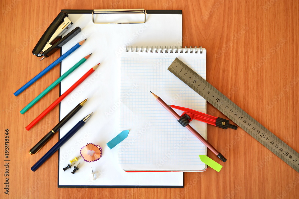 Cell notepad for writing with ring, compass, ruler, pins, color pencils, fountain ballpoint pen, clipboard, black stapler, mark and shaving on wooden table in office Cell notepad for writing with ring, compass, ruler, pins, color pencils, fountain ballpoint pen, clipboard, black stapler, mark and shaving on wooden table in office