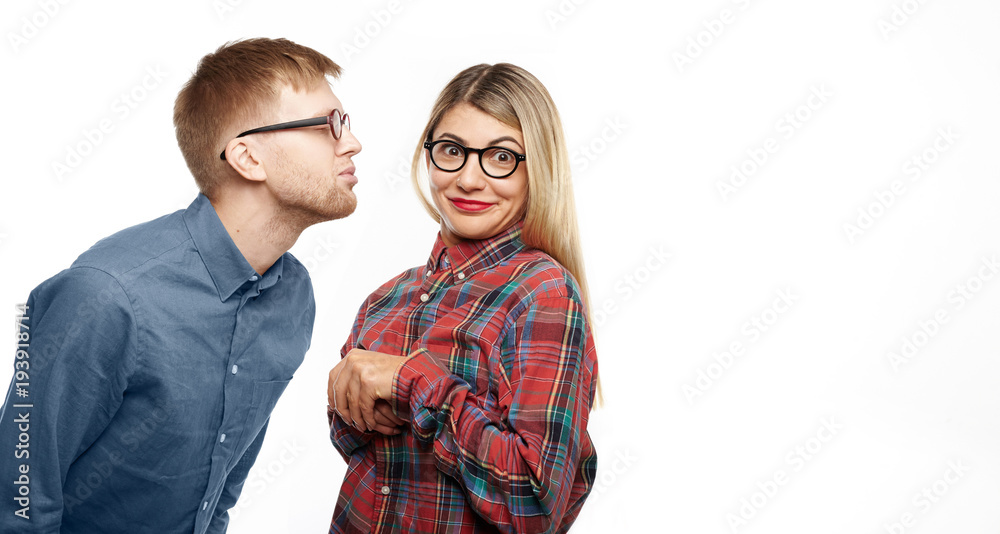 Horizontal shot of funny nerdy guy with stubble and eyeglasses closing eyes and pouting lips as if going to kiss attractive young woman who is feeling lost, looking at camera with confused smile Horizontal shot of funny nerdy guy with stubble and eyeglasses closing eyes and pouting lips as if going to kiss attractive young woman who is feeling lost, looking at camera with confused smile