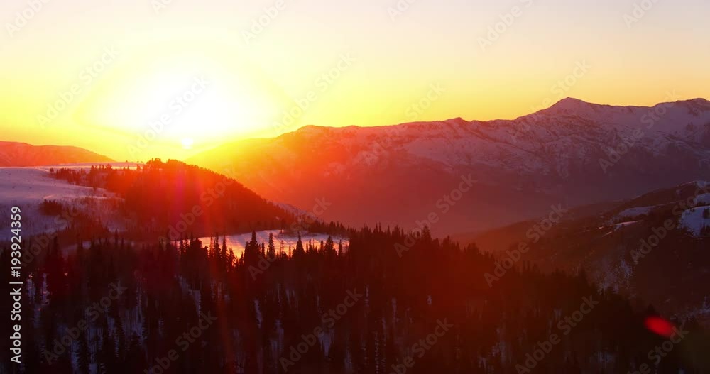 Aerial view flying over winter forest at sunset with snow covered mountains in background