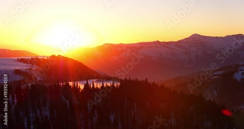 Aerial view flying over winter forest at sunset with snow covered mountains in background