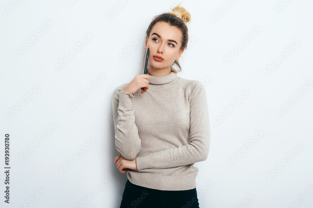 Portrait of Thinking woman with appealing smile, having hair bun in sweater isolated on white background holding pen and have a pensive face. Beautiful female showing emotions. People Beauty Fashion Portrait of Thinking woman with appealing smile, having hair bun in sweater isolated on white background holding pen and have a pensive face. Beautiful female showing emotions. People Beauty Fashion