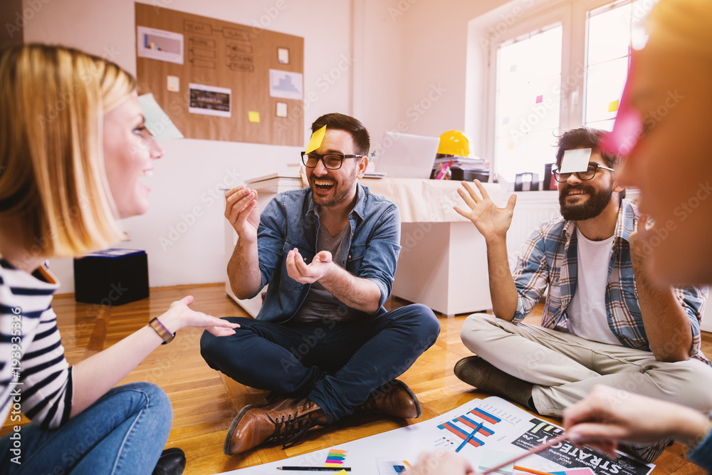 Φωτογραφία Pretty young designer coworkers enjoying free time and raising togetherness and team spirit while sitting in a circle on the floor of the office and playing games with paper stickers on the forehead