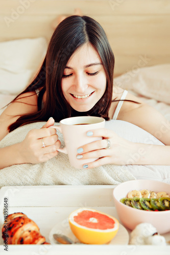Breakfast in bed / beautiful brunette woman holding white porcelain cup in hand and smiling lying in bed