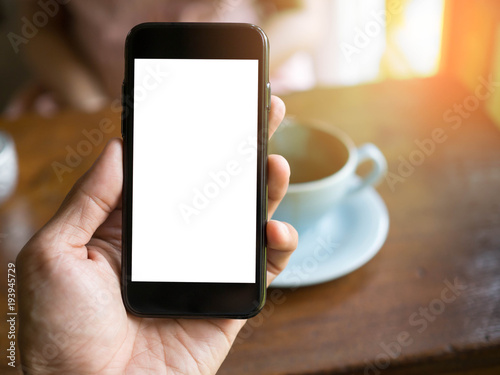 image of man's hand is holding a black cell phone with blank white screen and blur silver laptop on wood table in coffee cafe  background