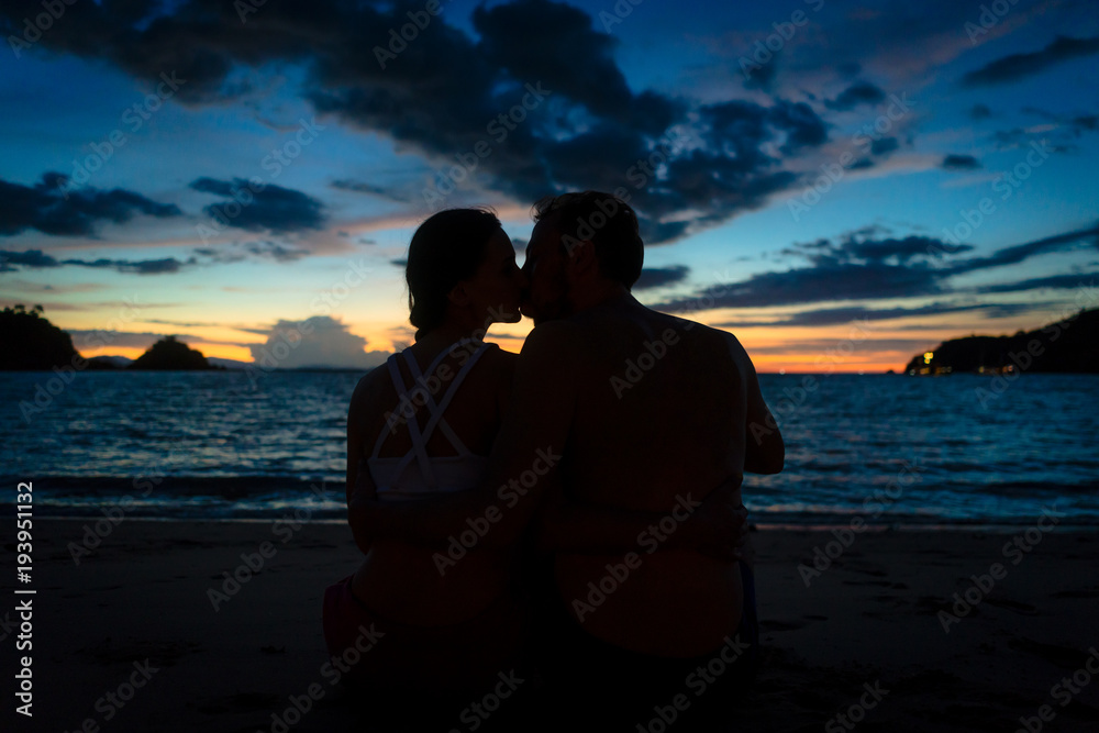 Rear view of a young couple kissing while sitting on a dreamy beach at dusk during summer vacation or honeymoon in Flores Island, Indonesia