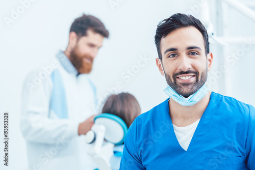 Portrait of a confident dental surgeon wearing blue protective uniform and surgical mask while looking at camera in a modern dental office with reliable specialists
